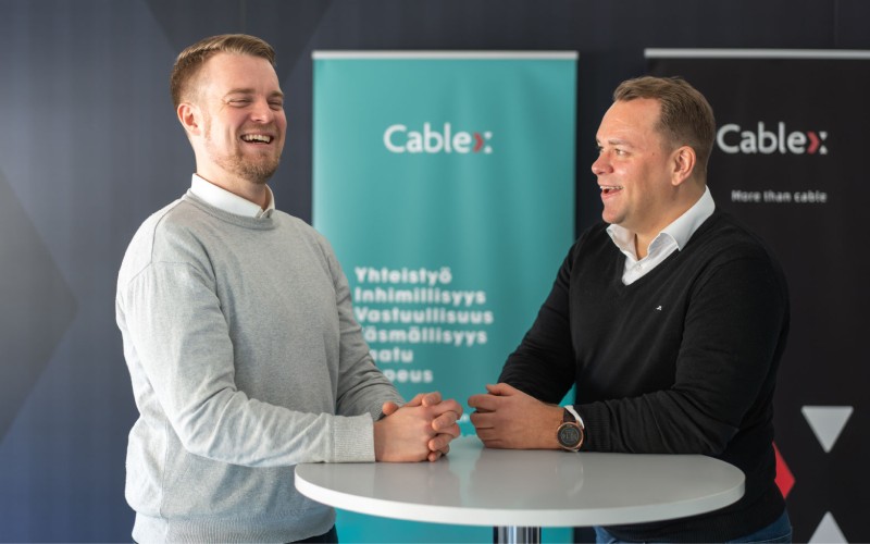 Kaks Cablex töötajat vestlevad ja naeravad koos laua ääres, taustal on näha Cablexi bännerid.